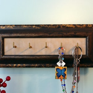 May include: A key holder made from a distressed, dark brown picture frame with gold accents. The frame has five brass hooks and is hanging on a light blue wall. Two keychains with colourful beads and charms are hanging from the hooks.