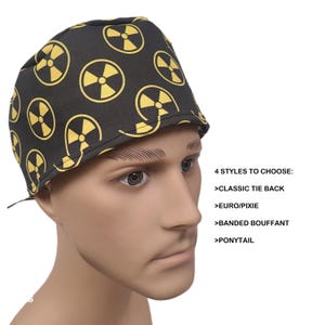May include: Black surgical scrub cap with a repeating yellow radiation symbol pattern. The cap is displayed on a mannequin head. Text on the right side of the image reads "4 STYLES TO CHOOSE: CLASSIC TIE BACK, EURO/PIXIE, BANDED BOUFFANT, PONYTAIL."