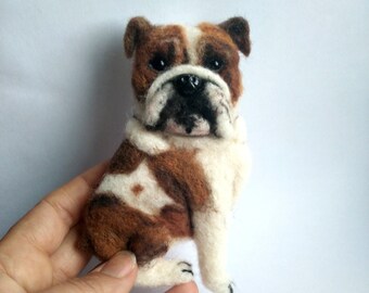 May include: A handmade felt bulldog figurine in a sitting position. The dog has brown and white fur with black accents on the nose and paws. The figurine is detailed and appears to be crafted with care.