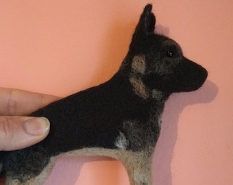 May include: A handmade felted German Shepherd dog figurine. The dog is black and tan, with detailed features. The figurine is posed in a standing position, with its ears perked up. The background is a solid pink color.