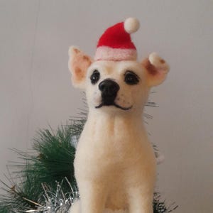 Può includere: Una statuina di cane bianco in feltro con un cappello da Babbo Natale rosso e bianco, seduta su un albero di Natale verde con orpelli argentati. Il cane ha occhi neri e naso nero. Decorazione natalizia festosa.