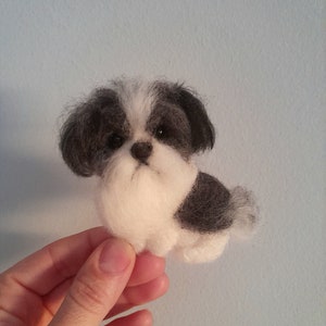 Pode incluir: Figura de cão Shih Tzu de feltro feita à mão. O cão pequeno tem uma pelagem preta e branca, com uma barba e peito brancos e fofos. O cão está sentado, com a cauda ligeiramente visível. O fundo é azul claro.
