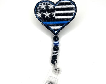 May include: A black and white heart-shaped badge holder with a blue line and stars. The holder is attached to a black and white beaded chain and a clear plastic badge clip.
