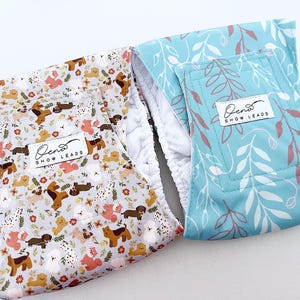 Puede incluir: Dos accesorios para mascotas con diseños de perros. Uno tiene un estampado de varias razas de perros y flores sobre un fondo gris claro. El otro es azul turquesa con un diseño de hojas blancas y granates. Ambos tienen interior blanco y etiqueta "Oeno SHOW LEADS."