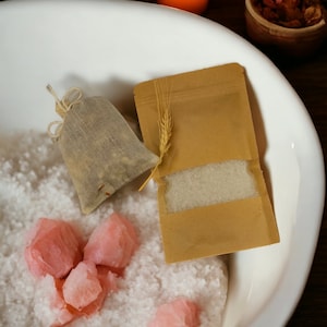 May include: A white bowl filled with white bath salts, pink bath salts, and a small brown pouch. A small brown paper bag with a wheat stalk design and a white powder inside is also in the bowl. A lit candle and a small brown bowl with dried herbs are in the background.