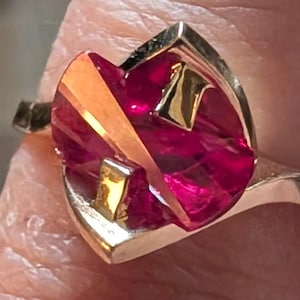 May include: A gold ring featuring a large, faceted, deep pinkish-red gemstone set within a polished gold frame. The metal band curves around the central stone, which has a textured, irregular surface with sharp angles and reflective gold accents.