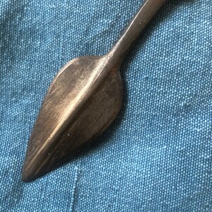 May include: A bronze-coloured metal arrowhead with a pointed tip and a flat, rounded base. The arrowhead is about 12.5 cm long.