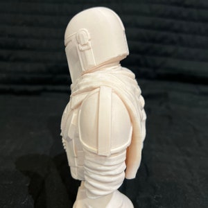 Puede incluir: Busto de resina pintado de blanco de un personaje mandaloriano de la franquicia Star Wars. El busto presenta un casco y una armadura detallados, y est&aacute; montado sobre una base redonda.