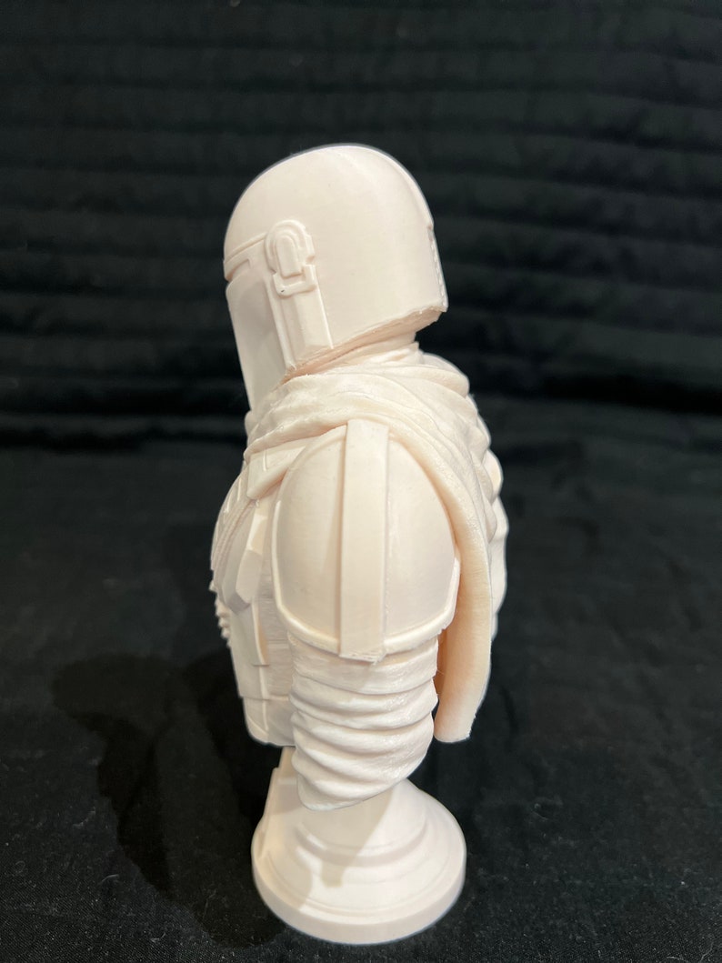 Puede incluir: Busto de resina pintado de blanco de un personaje mandaloriano de la franquicia Star Wars. El busto presenta un casco y una armadura detallados, y est&aacute; montado sobre una base redonda.