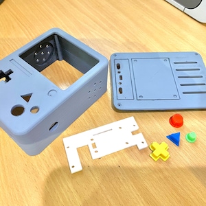 Könnte beinhalten: Ein 3D-gedrucktes graues Plastikgehäuse für eine Spielkonsole mit einer weißen Plastikplatine und bunten Knöpfen. Das Gehäuse hat eine Öffnung für den Bildschirm, Aussparungen für die Knöpfe und ein Loch für den Lautsprecher. Die Platine ist rechteckig mit Aussparungen für Bauteile. Die Knöpfe sind rot, gelb, grün und blau.