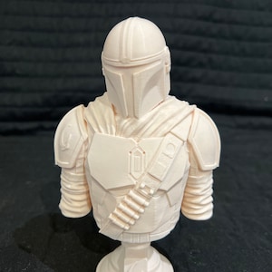Puede incluir: Un busto de pl&aacute;stico blanco de un personaje mandaloriano de la franquicia Star Wars. El busto lleva un casco y una armadura, y sostiene un rifle bl&aacute;ster. El busto est&aacute; sobre una base blanca.