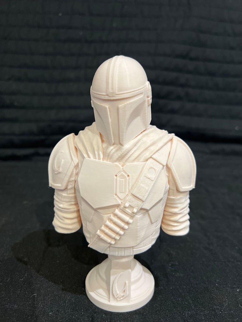 Puede incluir: Un busto de pl&aacute;stico blanco de un personaje mandaloriano de la franquicia Star Wars. El busto lleva un casco y una armadura, y sostiene un rifle bl&aacute;ster. El busto est&aacute; sobre una base blanca.