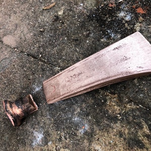 May include: A copper-colored axe head with a slightly curved blade and a rounded handle. The axe head is lying on a rough, gray surface.