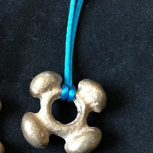 Puede incluir: Un colgante de color plateado con un diseño de estrella de cuatro puntas, colgando de un cordón turquesa.