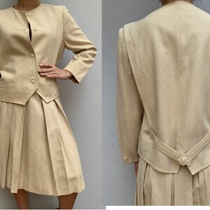 Puede incluir: Un traje de falda beige con una falda plisada y una chaqueta entallada. La chaqueta tiene un escote redondo y un cierre de bot&oacute;n.