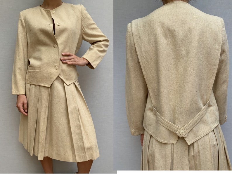 Puede incluir: Un traje de falda beige con una falda plisada y una chaqueta entallada. La chaqueta tiene un escote redondo y un cierre de bot&oacute;n.