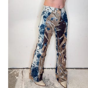 May include: Full-length denim pants featuring a patchwork design with a blue and white tie-dye pattern on one side and a brown, tan, and black feather-print fabric on the other. The pants have frayed, raw-edge seams and a button-fly closure.