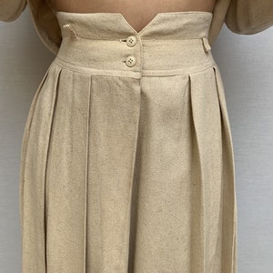 Puede incluir: Un pantal&oacute;n de cintura alta con pliegues en color beige claro. El pantal&oacute;n tiene cierre de bot&oacute;n y cremallera en la parte delantera y una cintura con trabillas.