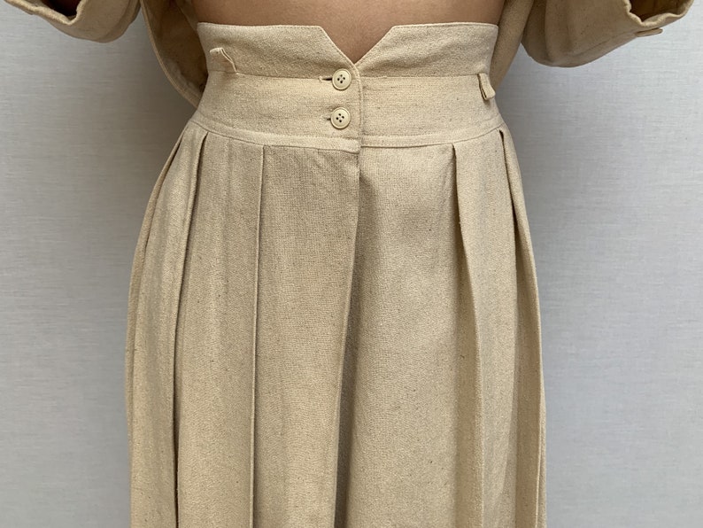Puede incluir: Un pantal&oacute;n de cintura alta con pliegues en color beige claro. El pantal&oacute;n tiene cierre de bot&oacute;n y cremallera en la parte delantera y una cintura con trabillas.