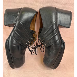 May include: A pair of black leather lace-up shoes with a thick block heel and a platform sole. The shoes feature stitched panel detailing on the sides, silver-toned metal eyelets for the black laces, and a tan interior lining visible at the opening.