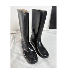 Puede incluir: Un par de botas de lluvia altas de color negro brillante con un acabado exterior liso y sin costuras, puntera redondeada y una caña estructurada hasta la pantorrilla. Las botas tienen una suela negra sólida y un diseño limpio.