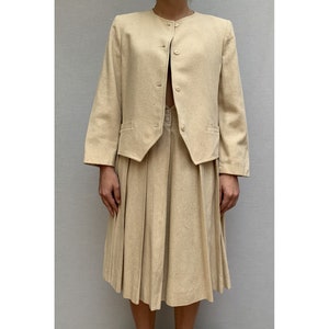 Puede incluir: Un traje de falda beige con una chaqueta de manga larga y una falda plisada.