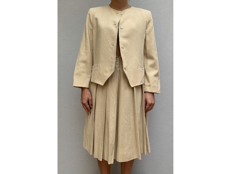 Puede incluir: Un traje de falda beige con una chaqueta de manga larga y una falda plisada.