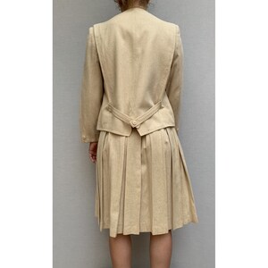 Puede incluir: Un traje de falda beige con una falda plisada y una chaqueta entallada. La chaqueta tiene un cierre de bot&oacute;n &uacute;nico en la espalda.