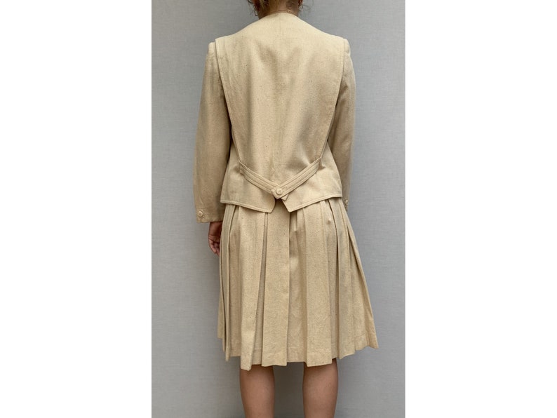 Puede incluir: Un traje de falda beige con una falda plisada y una chaqueta entallada. La chaqueta tiene un cierre de bot&oacute;n &uacute;nico en la espalda.