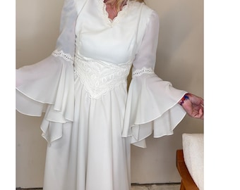 Puede incluir: Un vestido blanco hasta el suelo con mangas largas acampanadas en niveles y ribete de encaje en los codos. El corpiño incluye un escote en V con borde de encaje y un panel de encaje decorativo ancho en la cintura. La falda cae desde la cintura.