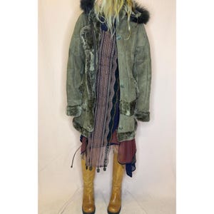 May include: An olive green suede coat with a dark brown faux fur lining, cuffs, and a hood trim. The coat features a button-front closure. Underneath is a layered dress with a dark patterned panel and a sheer burgundy hem, paired with tan boots.