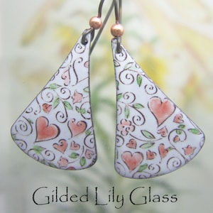 Op de afbeelding: Witte, driehoekige oorbellen met een hart- en bloemmotief. De oorbellen hebben een patroon van perzikkleurige harten, groene bladeren en kleine roze bloemen. De tekst "Gilded Lily Glass" staat onderaan.