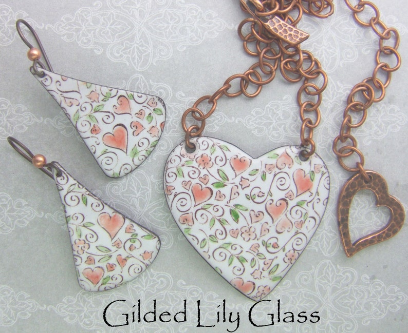 Op de afbeelding: Een sieradenset met een hartvormige hangerketting en bijpassende oorbellen. De witte emaille stukken zijn versierd met een patroon van perzikkleurige harten, groene bladeren en krullen. Het metaal heeft een koperkleur. De tekst "Gilded Lily Glass" staat onderaan.