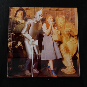May include: A vintage image of the characters from the Wizard of Oz. Dorothy, the Scarecrow, the Tin Man, and the Cowardly Lion are standing together in front of a brick wall. Dorothy is wearing a blue and white dress and red shoes. The Scarecrow is wearing a straw hat and a brown suit. The Tin Man is wearing a silver suit and a silver hat. The Cowardly Lion is wearing a brown fur coat and a brown mane.