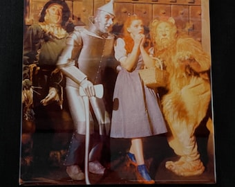 May include: A vintage image of the characters from the Wizard of Oz. Dorothy, the Scarecrow, the Tin Man, and the Cowardly Lion are standing together in front of a brick wall. Dorothy is wearing a blue and white dress and red shoes. The Scarecrow is wearing a straw hat and a brown suit. The Tin Man is wearing a silver suit and a silver hat. The Cowardly Lion is wearing a brown fur coat and a brown mane.