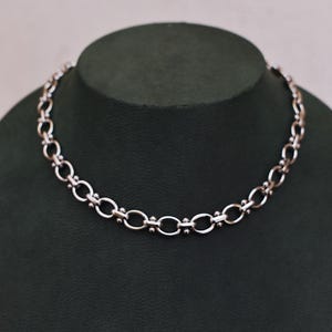 May include: A silver necklace with a chain-link design. The necklace is displayed on a dark green mannequin. The chain links alternate between oval and circular shapes, with small bead accents.