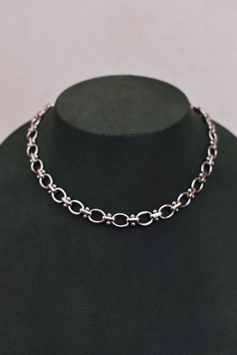 May include: A silver necklace with a chain-link design. The necklace is displayed on a dark green mannequin. The chain links alternate between oval and circular shapes, with small bead accents.