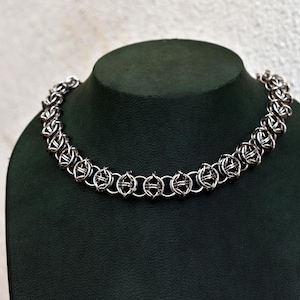 May include: A silver chainmail necklace displayed on a dark green jewelry bust. The necklace features an intricate design of interlocking metal rings, forming a textured, circular pattern. The necklace is designed to be worn around the neck.