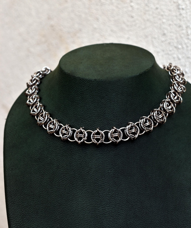 May include: A silver chainmail necklace displayed on a dark green jewelry bust. The necklace features an intricate design of interlocking metal rings, forming a textured, circular pattern. The necklace is designed to be worn around the neck.