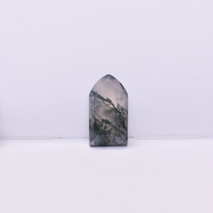 May include: A polished moss agate stone shaped like a small arch or tombstone. The stone features a translucent, milky white base with intricate, dark green, fern-like dendritic inclusions spread throughout the surface in a natural pattern.