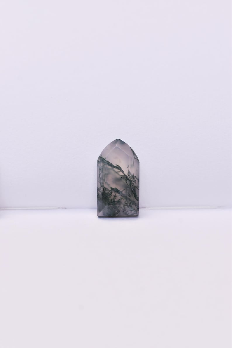 May include: A polished moss agate stone shaped like a small arch or tombstone. The stone features a translucent, milky white base with intricate, dark green, fern-like dendritic inclusions spread throughout the surface in a natural pattern.