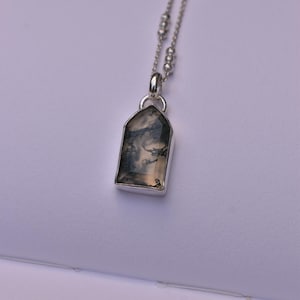 May include: A silver necklace featuring a pendant with a translucent, shield-shaped stone containing dark, branch-like inclusions. The pendant hangs from a delicate silver chain accented with small, evenly spaced silver beads along the length.