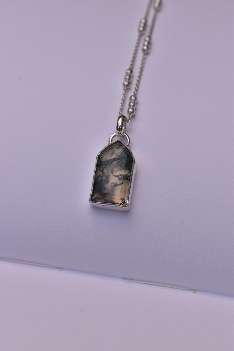 May include: A silver necklace featuring a pendant with a translucent, shield-shaped stone containing dark, branch-like inclusions. The pendant hangs from a delicate silver chain accented with small, evenly spaced silver beads along the length.