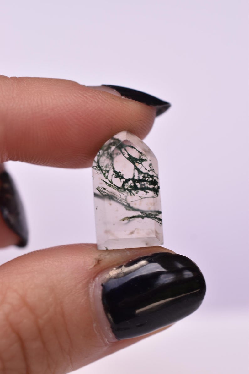 May include: A small, polished, clear quartz crystal tower with a pointed top and a flat base. The stone features distinct, dark green, moss-like dendritic inclusions that branch throughout the transparent interior of the mineral specimen.