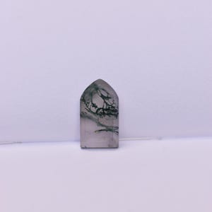 May include: A polished, arch-shaped moss agate gemstone featuring a translucent, light gray base with distinct, dark green, branch-like dendritic inclusions spreading throughout the stone, creating a natural, organic pattern within the crystal.