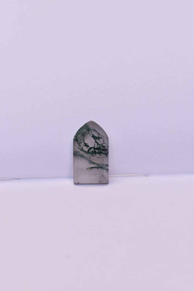 May include: A polished, arch-shaped moss agate gemstone featuring a translucent, light gray base with distinct, dark green, branch-like dendritic inclusions spreading throughout the stone, creating a natural, organic pattern within the crystal.