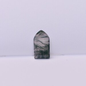 May include: A polished, semi-translucent stone cabochon shaped like a pointed arch or cathedral window. The stone features a clear, pale base with dark, moss-like inclusions that form intricate, branching, fern-like patterns throughout the interior.