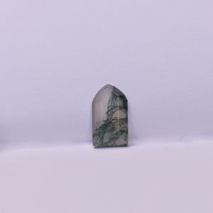 May include: A polished moss agate gemstone shaped like a small tombstone or arch, featuring a translucent, milky white base with intricate, dark green, fern-like dendrite inclusions spreading throughout the stone's surface in a natural pattern.