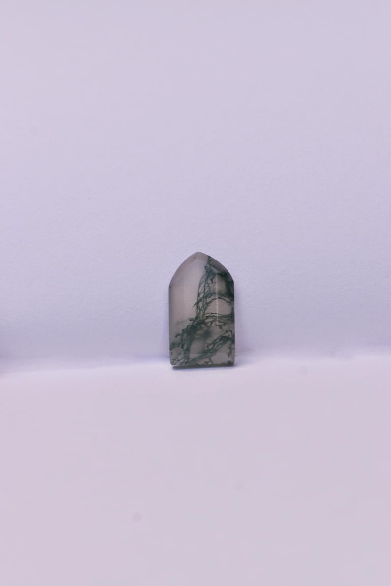 May include: A polished moss agate gemstone shaped like a small tombstone or arch, featuring a translucent, milky white base with intricate, dark green, fern-like dendrite inclusions spreading throughout the stone's surface in a natural pattern.