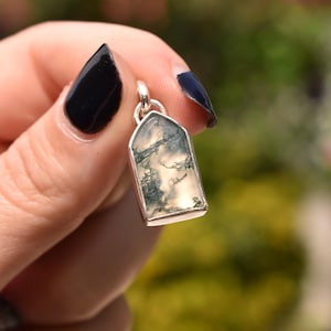 May include: A pendant featuring a translucent, shield-shaped moss agate stone with dark green, fern-like inclusions. The stone is encased in a polished silver-colored metal bezel setting with a small circular bail at the top for attachment.